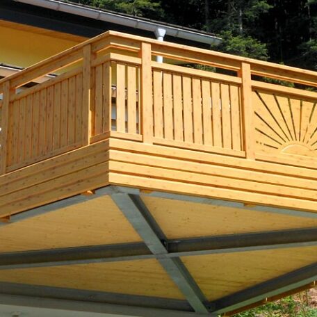 Holzbalkon mit dekorativem Geländer und Sonnenmuster, angebracht an einem modernen Haus. de HANSEL.kraus metalltechnik gmbh