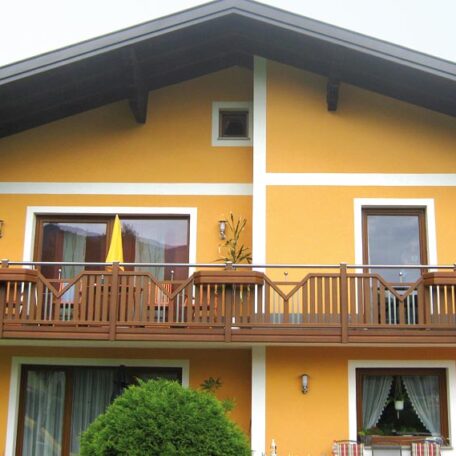 Gelbes Haus mit Holz-Balkon, weißen Fensterrahmen und grünen Pflanzen im Vordergrund. de HANSEL.kraus metalltechnik gmbh