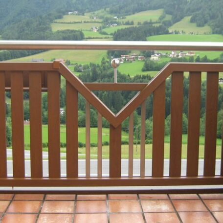 Holzgeländer mit Ziermuster auf Balkon, dahinter grüne Wiesen und Berge im Hintergrund. HANSEL.kraus metalltechnik gmbh