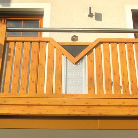 Holzbalkon mit geometrischem Muster vor hellgelber Hauswand und Tür, Tageslicht. HANSEL.kraus metalltechnik gmbh