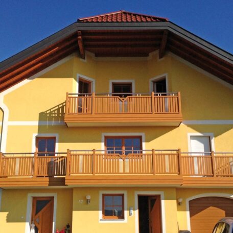 Gelbes Einfamilienhaus mit Holz-Balkonen und rotem Dach unter blauem Himmel. HANSEL.kraus metalltechnik gmbh