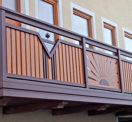 Holzbalkon mit geschnitztem Sonnenmotiv vor gelber Hausfassade und mehreren Fenstern, Freiburg. HANSEL.kraus metalltechnik gmbh