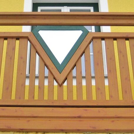 Holzbalkon mit dreieckigem Zierelement vor gelber Hausfassade und einem Fenster im Hintergrund. HANSEL.kraus metalltechnik gmbh