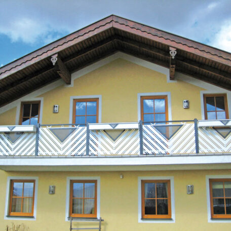 Gelbes Haus mit Holzveranda, weißen Balkongeländern und mehreren Fenstern unter blauem Himmel. HANSEL.kraus metalltechnik gmbh
