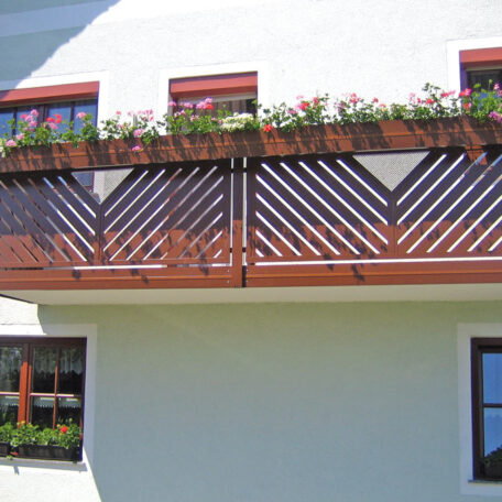 Holzbalkon mit Blumen in Kästen an hellgrüner Hausfassade, darunter Fenster mit Blumendekor. HANSEL.kraus metalltechnik gmbh