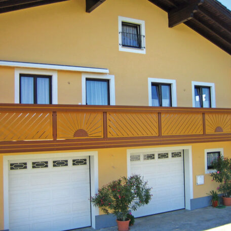 Zweistöckiges gelbes Haus mit Balkon, weißen Fenstern und zwei Garagentoren, davor Kübelpflanzen. HANSEL.kraus metalltechnik gmbh