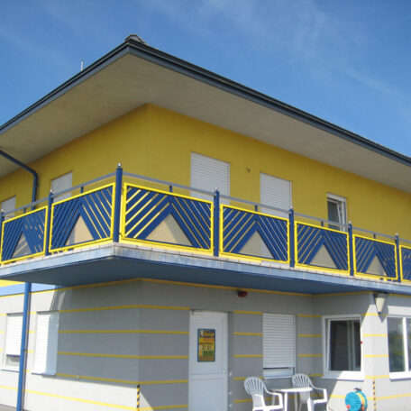 Zweistöckiges gelb-blaues Gebäude mit Balkon und Sonnenmuster, blauer Himmel. de HANSEL.kraus metalltechnik gmbh