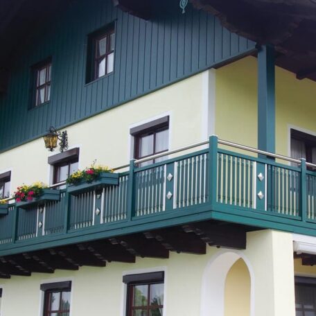 Grünes Holzhaus mit Balkon, Blumenkästen und weißen Fensterrahmen an der Fassade. HANSEL.kraus metalltechnik gmbh