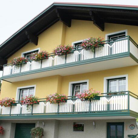 Gelbes Haus mit Blumenkästen an allen Balkonen vor blauem Himmel, Frontalansicht. HANSEL.kraus metalltechnik gmbh