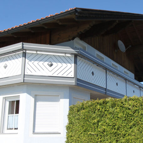 Zweigeschossiges Haus mit weißem Holzbalkon und schrägem Dach hinter einer grünen Hecke. HANSEL.kraus metalltechnik gmbh