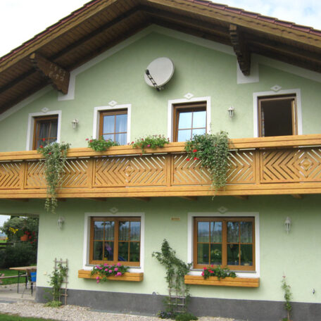 Zweistöckiges Haus mit grünem Putz, Holzbalkon und Blumenkästen an den Fenstern. HANSEL.kraus metalltechnik gmbh