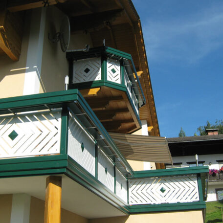 Detailreich verzierter Balkon mit grün-weißen Holzgeländern an einem traditionellen Haus, blauer Himmel. HANSEL.kraus metalltechnik gmbh