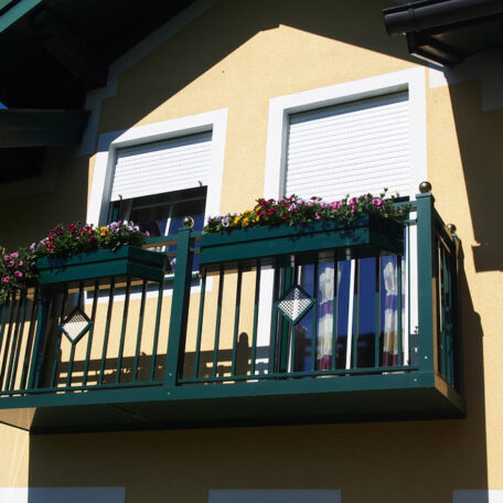 Blumengeschmückter Balkon an gelbem Haus mit zwei geschlossenen weißen Fenstern, Sonnenschein. HANSEL.kraus metalltechnik gmbh