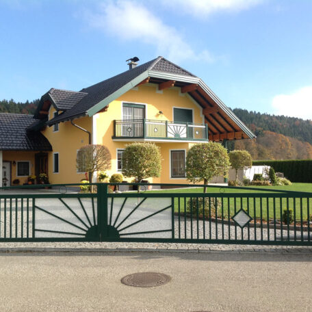 Gelbes Einfamilienhaus mit Balkon, grünem Zaun und gepflegtem Vorgarten an sonnigem Tag. HANSEL.kraus metalltechnik gmbh