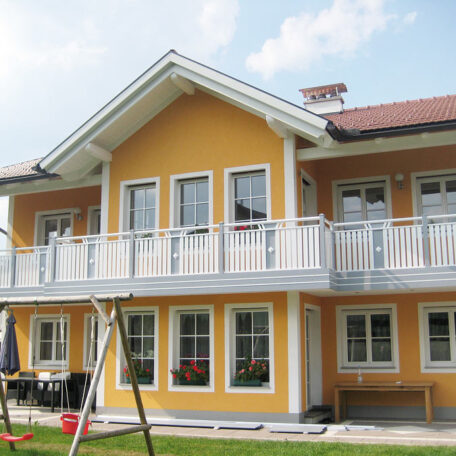 Zweistöckiges gelbes Einfamilienhaus mit Balkon, weißem Zaun und Garten im Vordergrund. HANSEL.kraus metalltechnik gmbh