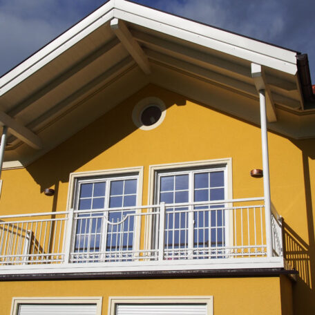 Gelbes Haus mit weißem Balkon und großen Fenstern unter einem Spitzdach bei Tageslicht. HANSEL.kraus metalltechnik gmbh