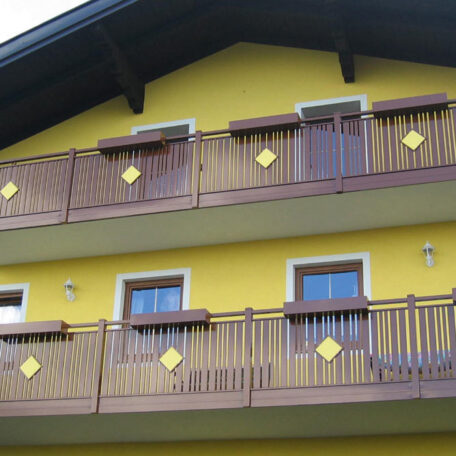 Zweistöckiges gelbes Haus mit braunen Holzbalkonen und weißen Fensterrahmen, Vorderansicht. HANSEL.kraus metalltechnik gmbh