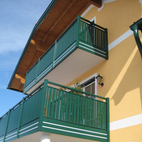 Zweistöckiges Wohnhaus mit gelber Fassade und grünen Metallbalkonen vor blauem Himmel. HANSEL.kraus metalltechnik gmbh