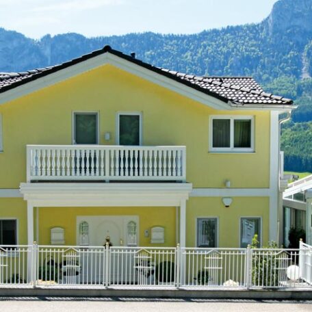 Zweistöckiges gelbes Haus mit Balkon, weißem Zaun und Bergen im Hintergrund. de HANSEL.kraus metalltechnik gmbh
