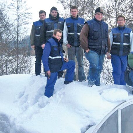 Sechs Männer in Winterkleidung stehen gemeinsam im Schnee auf einem Dach, Bäume im Hintergrund. de HANSEL.kraus metalltechnik gmbh