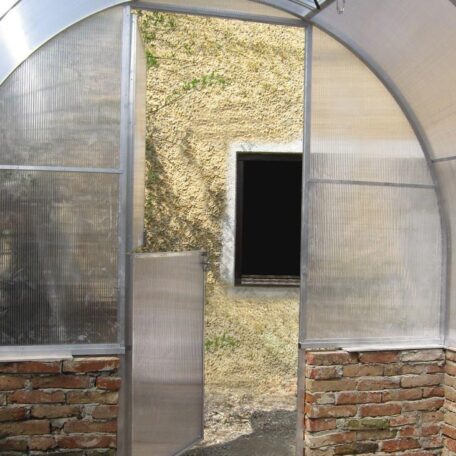 Ein transparentes Gewächshaus mit geöffneter Tür steht vor einer alten Steinmauer. de HANSEL.kraus metalltechnik gmbh