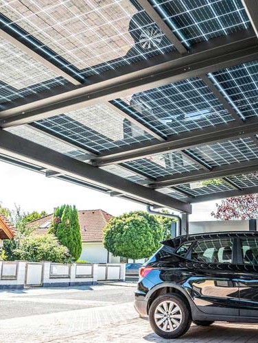 Carport mit Solardach neben einem geparkten Auto, im Hintergrund Wohnhäuser und grüne Bäume. HANSEL.kraus metalltechnik gmbh