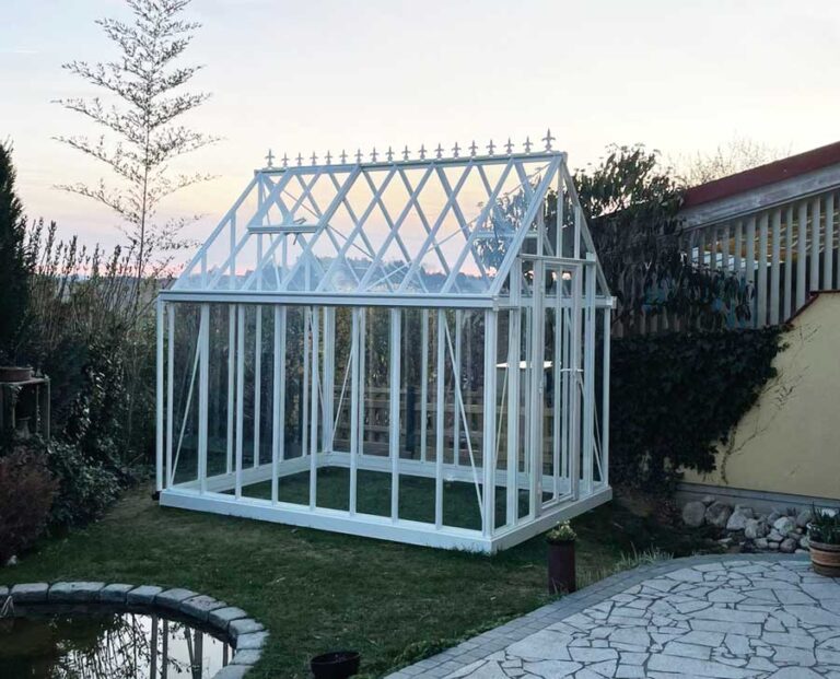 Weißes Gewächshaus aus Glas steht im Garten neben einem kleinen Teich bei Sonnenuntergang. HANSEL.kraus metalltechnik gmbh