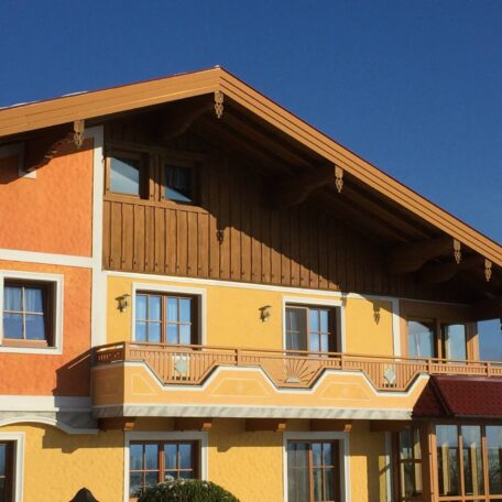 Traditionelles, gelb-orange gestrichenes Haus mit Holzverkleidung und Balkon vor blauem Himmel. HANSEL.kraus metalltechnik gmbh