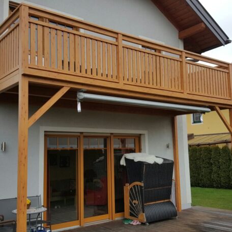 Holzbalkon mit Geländer an Einfamilienhaus, darunter Terrasse mit Gartenmöbeln, Sicht auf Garten. de HANSEL.kraus metalltechnik gmbh