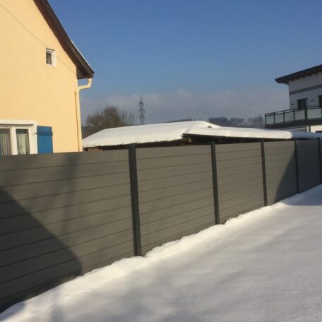 Grauer Sichtschutzzaun im Schnee zwischen zwei Wohnhäusern unter blauem Himmel. HANSEL.kraus metalltechnik gmbh