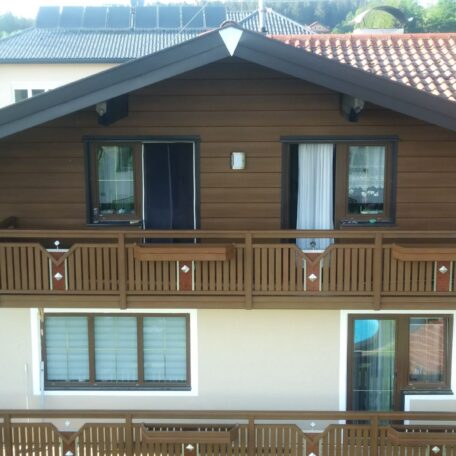Zweistöckiges Holzhaus mit Balkon, geöffneten Fenstern und Vorhängen vor sonnigem Hintergrund. HANSEL.kraus metalltechnik gmbh