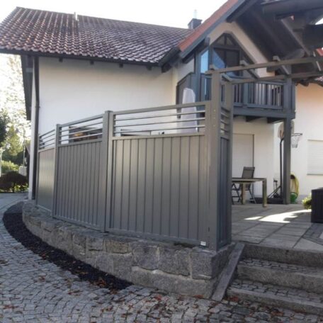 Ein modernes Haus mit grauem Metallzaun, gepflastertem Weg und überdachter Terrasse. de HANSEL.kraus metalltechnik gmbh
