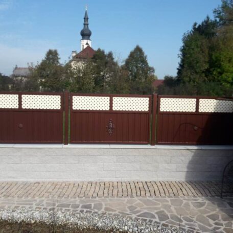 Braunes Hoftor vor Kopfsteinpflaster, dahinter Bäume und eine Kirche mit Zwiebelturm im Hintergrund. de HANSEL.kraus metalltechnik gmbh