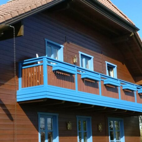 Traditionelles Holzhaus mit blauem Balkon, sonnige Fassade, zwei Fenster im Obergeschoss. HANSEL.kraus metalltechnik gmbh