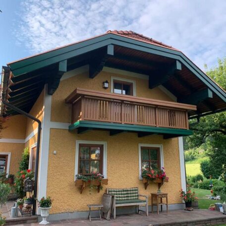Gemütliches gelbes Haus mit Holz-Balkon, Blumen und grünem Garten im Sommer. HANSEL.kraus metalltechnik gmbh