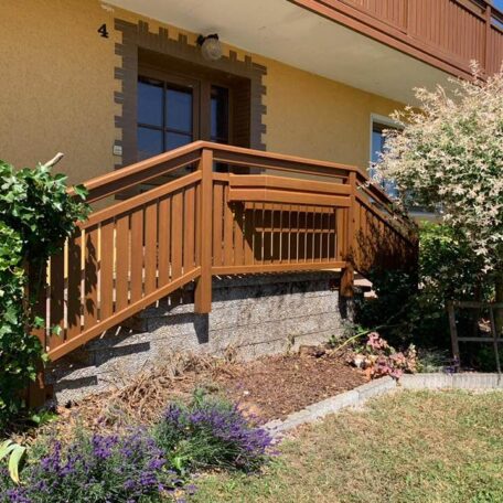 Holzgeländer auf Terrasse eines Einfamilienhauses, umgeben von Garten und blühenden Pflanzen. HANSEL.kraus metalltechnik gmbh