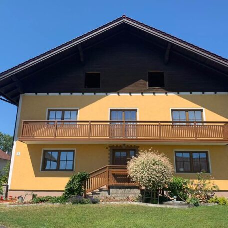 Gelbes Einfamilienhaus mit Holzbalkon, Satteldach und gepflegtem Garten bei Sonnenschein. HANSEL.kraus metalltechnik gmbh