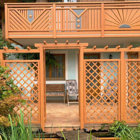 Holzveranda mit Rankgittern und Sitzbank vor einem modernen Haus, umgeben von Gartenpflanzen. HANSEL.kraus metalltechnik gmbh