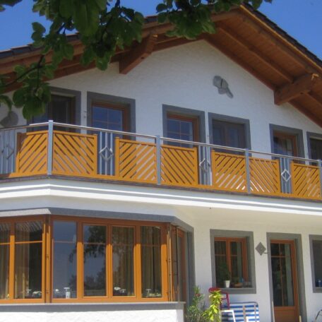 Zweistöckiges Haus mit Holzbalkon, großen Fenstern und Satteldach bei sonnigem Wetter. HANSEL.kraus metalltechnik gmbh