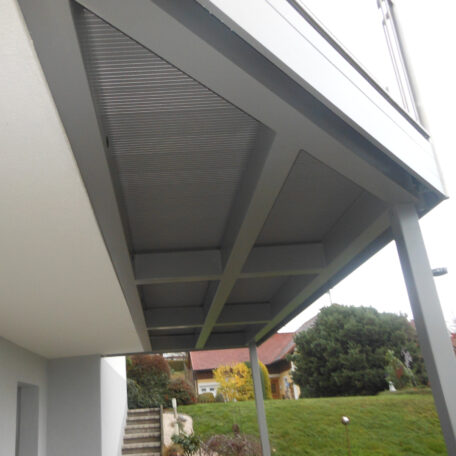 Unter einem modernen Balkon führt eine Treppe zum Garten mit Wiese und Häusern im Hintergrund. HANSEL.kraus metalltechnik gmbh