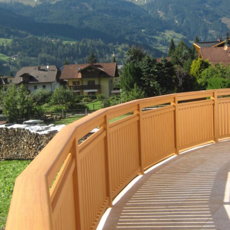 Holzgeländer mit Bergblick und traditionellen Häusern im Hintergrund bei sonnigem Wetter. HANSEL.kraus metalltechnik gmbh