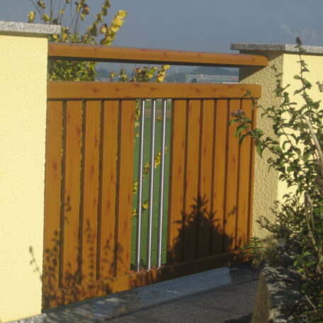 Holztor mit senkrechten Latten und Ziermetall zwischen zwei gelben Mauern im Sonnenlicht. HANSEL.kraus metalltechnik gmbh