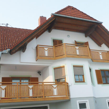 Zweistöckiges Haus mit Holzbalkonen, weißen Wänden und rotem Satteldach bei Tag. HANSEL.kraus metalltechnik gmbh