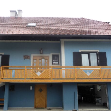 Zweistöckiges blaues Haus mit rotem Satteldach, Holzgeländer und Balkon, ein Carport darunter. HANSEL.kraus metalltechnik gmbh