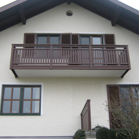 Zweistöckiges Haus mit braunem Holzbalkon, Fenstern und Eingangstreppe; Außenansicht bei Tageslicht. HANSEL.kraus metalltechnik gmbh
