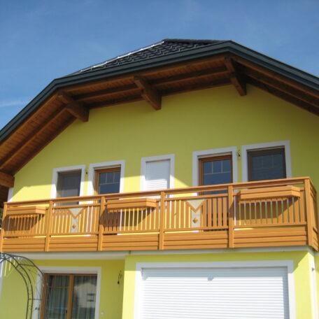 Gelbes Einfamilienhaus mit Holz-Balkon und weißer Haustür unter blauem Himmel. HANSEL.kraus metalltechnik gmbh