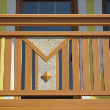 Holzgeländer mit modernem, geometrischem Muster auf einem Balkon, gelbes Haus im Hintergrund. HANSEL.kraus metalltechnik gmbh