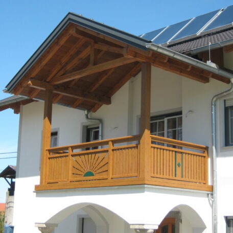 Holzbalkon mit Sonnenmotiv an weißem Haus, darüber Satteldach und Solarpanels bei Sonnenschein. HANSEL.kraus metalltechnik gmbh