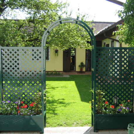 Garten mit grünem Tor, Rasenfläche, Blumenbeeten und gelbem Haus im Hintergrund sichtbar. HANSEL.kraus metalltechnik gmbh