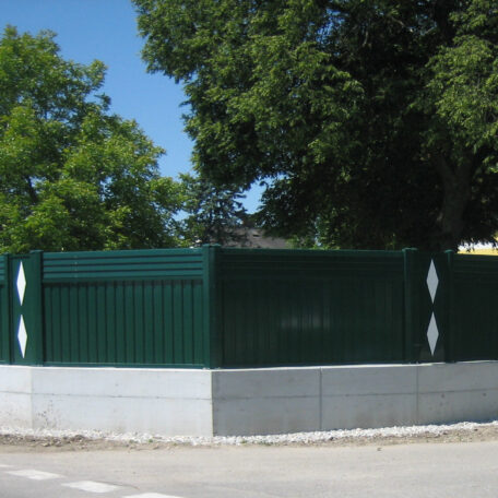 Grüner Metallzaun auf Betonsockel an Straßenecke, umgeben von Bäumen und Gehweg. HANSEL.kraus metalltechnik gmbh